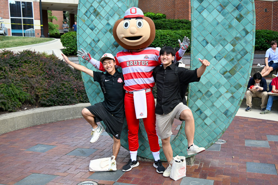 Brutus Buckeye, the Ohio State University mascot, poses with two people in front of a large green arch sculpture.